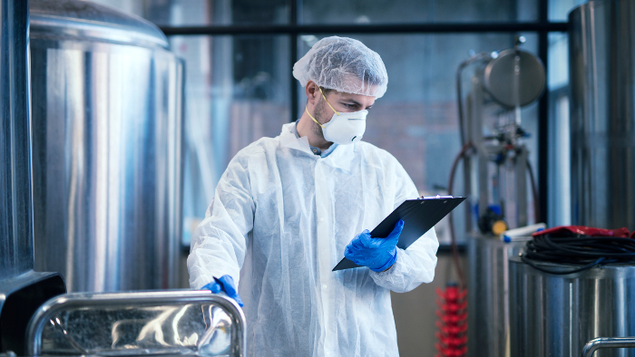 Worker in protective gear holding a clipboard in a facility with industrial equipment, emphasizing the role of industrial compressors in sectors like food and beverage processing.